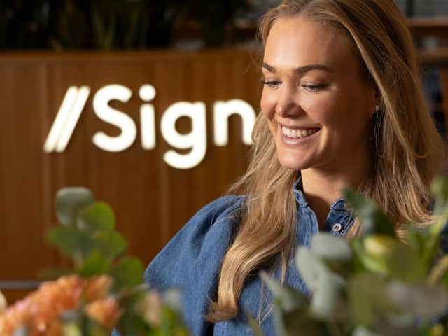 Ung blond kvinna i förgrunden och i bakgrunden står en servicedesk med skylten sign