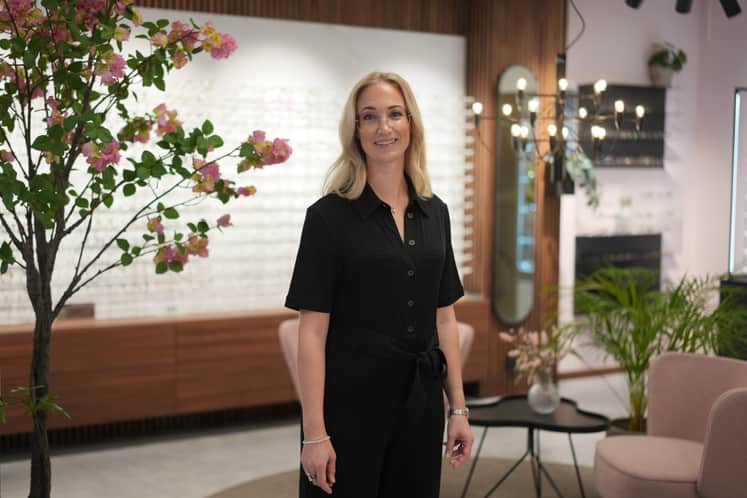 Medelålders kvinna klädd i svart lång dress, står mitt i butiken och man ser glasögon på väggen bakom