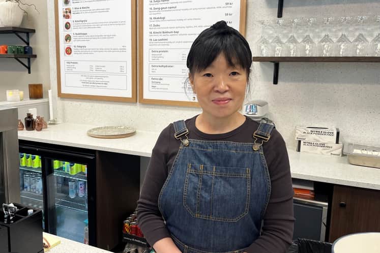 Person står bakom disken i ett modernt café med ljus inredning. I bakgrunden syns meny­tavlor, hyllor med glas och en kyl med drycker, vilket skapar en varm och välkomnande cafémiljö.