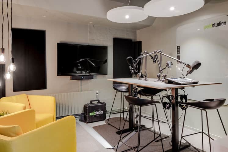 Modern podcaststudio med professionell inspelningsutrustning, mikrofoner och högt bord med barstolar. Ljusa väggar, stora taklampor och gula fåtöljer skapar en stilren och kreativ miljö för samtal och inspelning