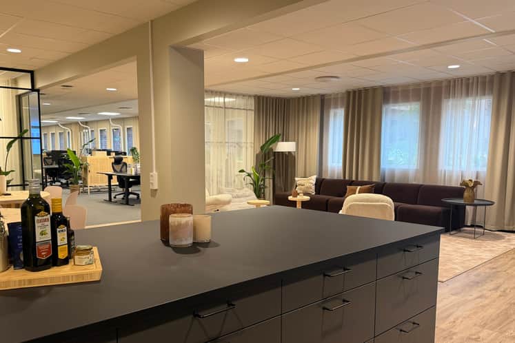 Modernt och stilrent kontorslandskap med öppen planlösning. I förgrunden syns en mörk köksö med dekorativa ljus och olivoljor, och i bakgrunden finns en loungehörna med mörk soffa, små bord, växter och ljusa gardiner som skapar en varm och inbjudande atmosfär.