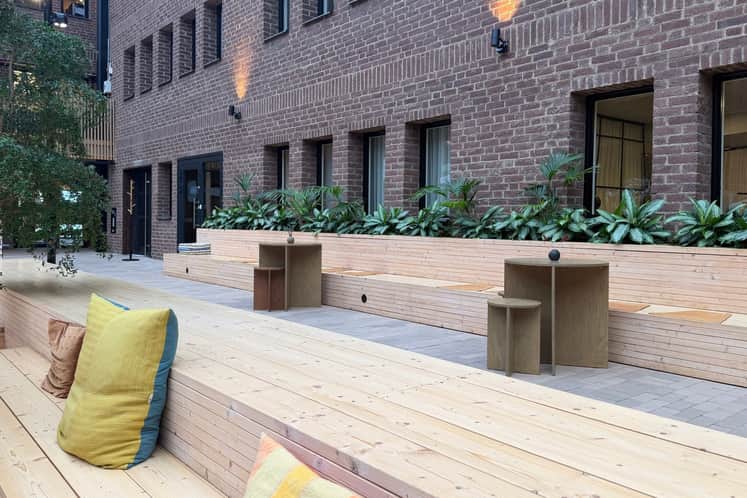 Innergård med modern och inbjudande uteplats intill en tegelbyggnad. Långa sittbänkar i ljust trä, planteringar med gröna växter och mindre bord skapar en lugn och social miljö mellan byggnaderna.