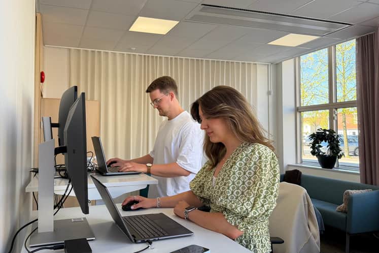 Två personer arbetar vid höj- och sänkbara skrivbord i ett ljust och modernt kontor. En kvinna sitter vid en dator i förgrunden medan en man står och arbetar vid ett skrivbord bakom henne. Stora fönster och ljusa material skapar en öppen och trivsam arbetsmiljö.