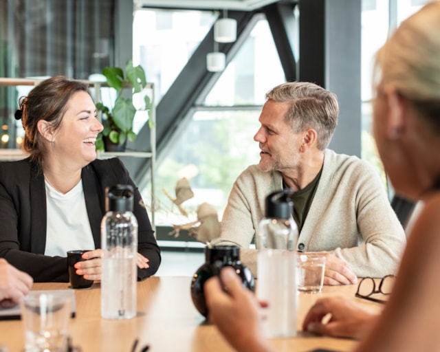 Tre personer sitter runt ett bord på ett kontor, leendes.