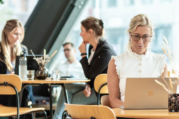 En kvinna i vitt linne, glasögon och ljust hår sitter och jobbar med en laptop