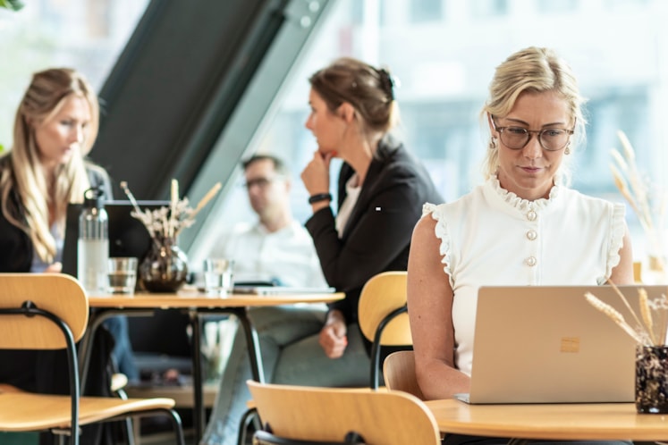 En kvinna sitter och jobbar vid sin laptop med människor i bakgrunden